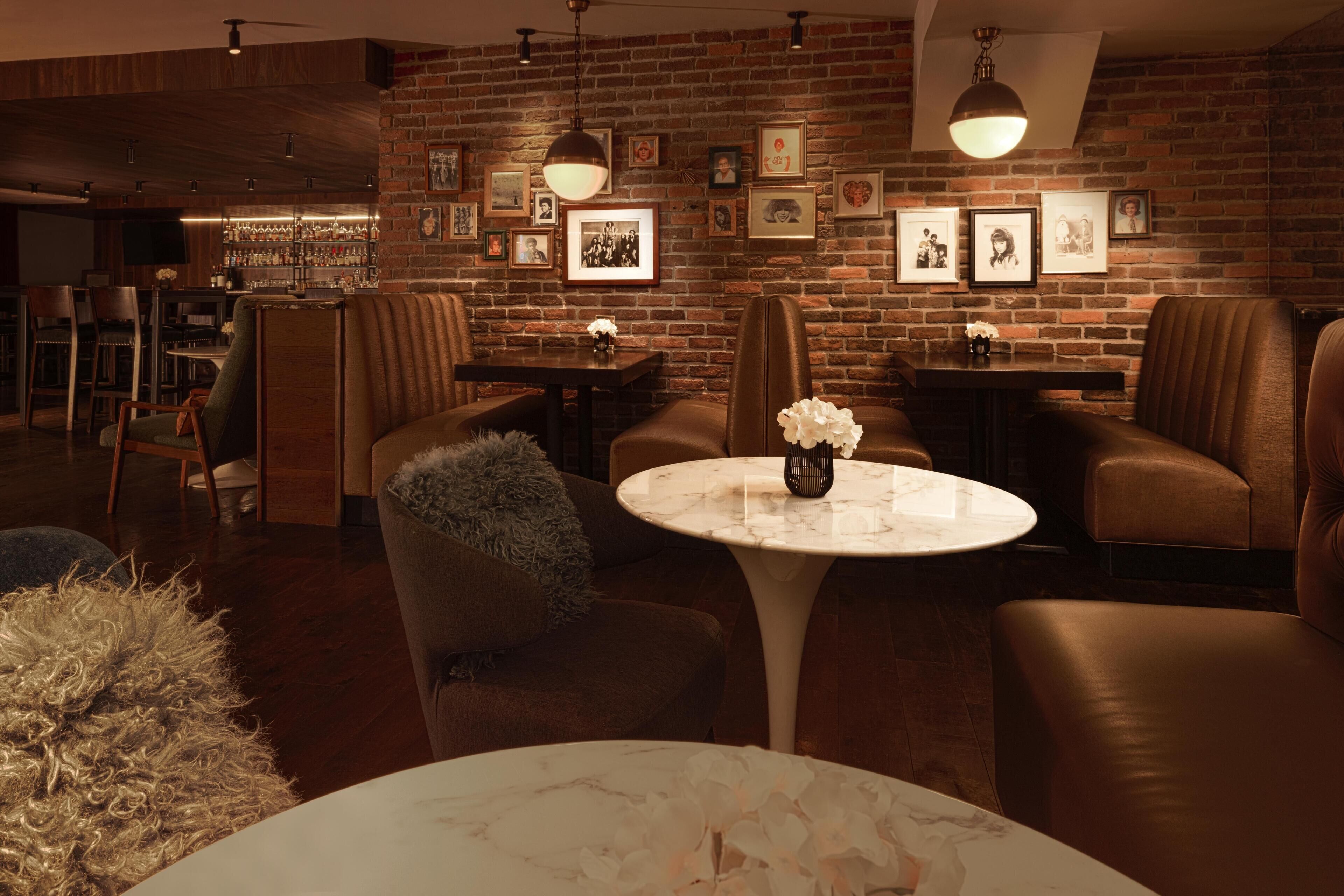 Un coin bar accueillant avec des sièges en cuir, une table ronde agrémentée d’un vase et un mur de briques orné de tableaux encadrés.