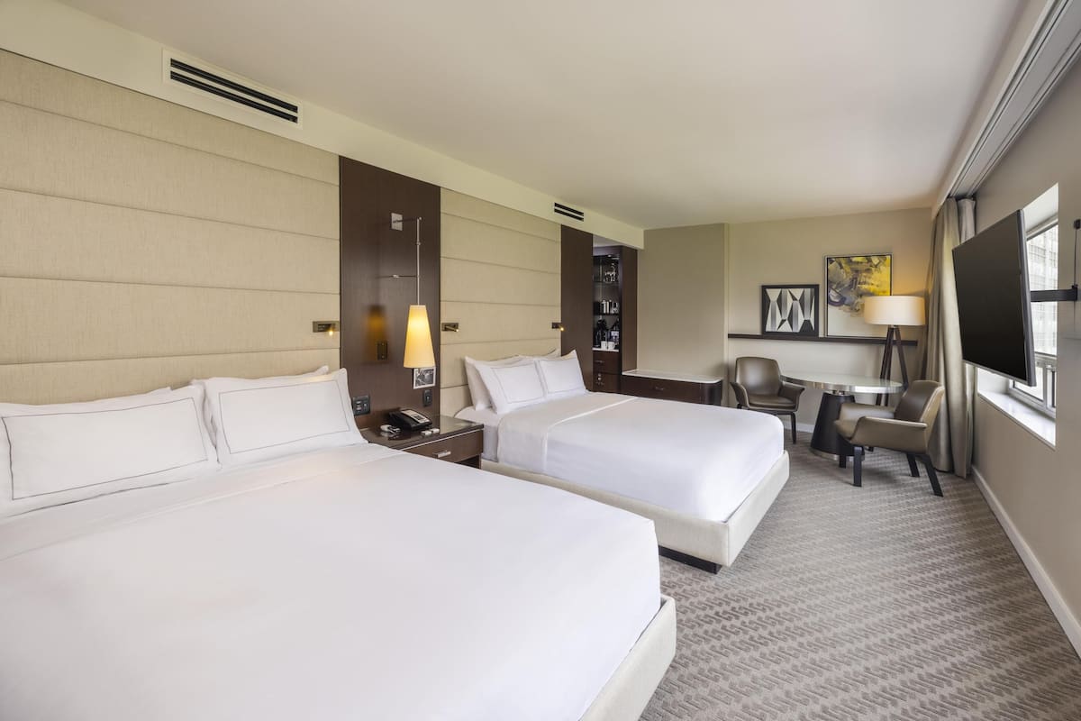 A hotel room with two beds, a desk, a chair, a TV, and a lamp.