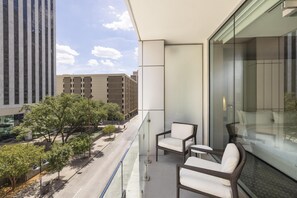 A balcony with wicker chairs, a glass railing, and a view of a city street and buildings.