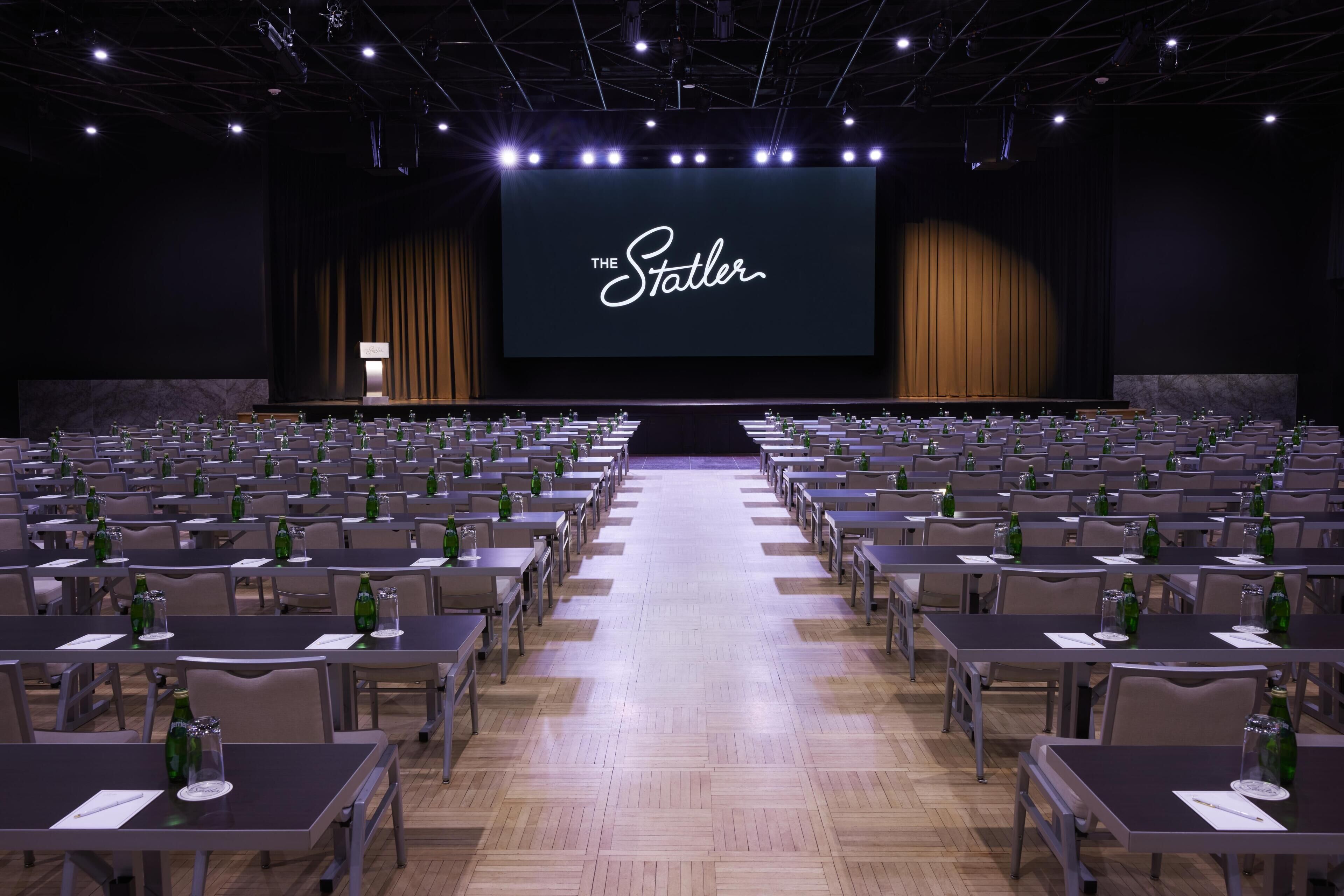 Un auditorio con filas de mesas, un escenario con una pantalla que muestra 'THE Statler' y un interior oscuro.