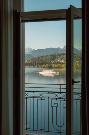 Blick durch eine Tür auf einen See mit einem Boot, dahinter Berge und ein Balkon mit Metallgeländer.