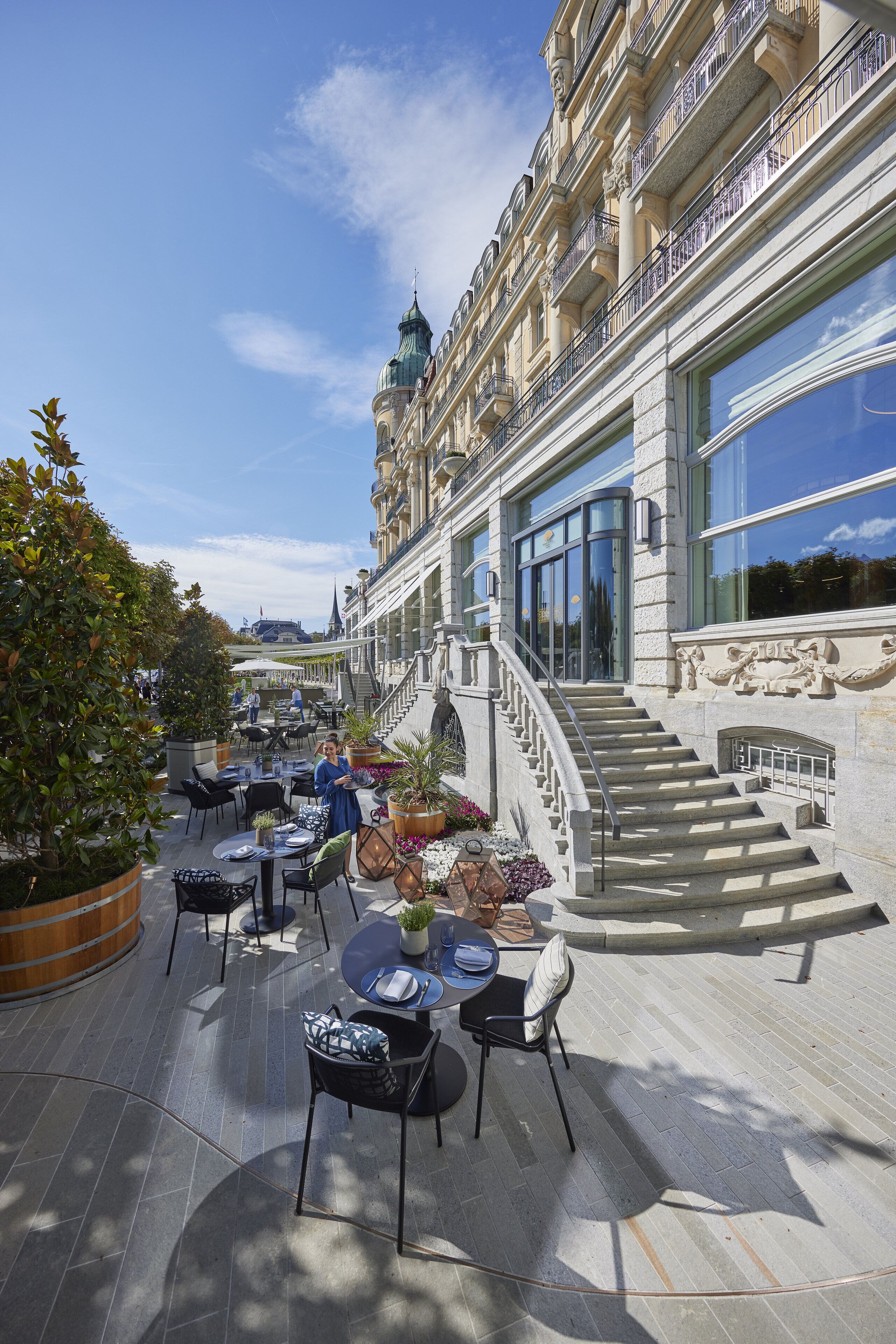 Un café al aire libre con mesas y sillas, una escalera y un edificio con ventanas amplias y una cúpula.