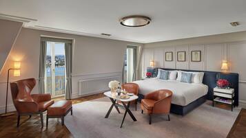 Una habitación de hotel moderna con una cama grande, dos sillones de cuero, una mesita redonda y vistas de la ciudad a través de amplios ventanales.