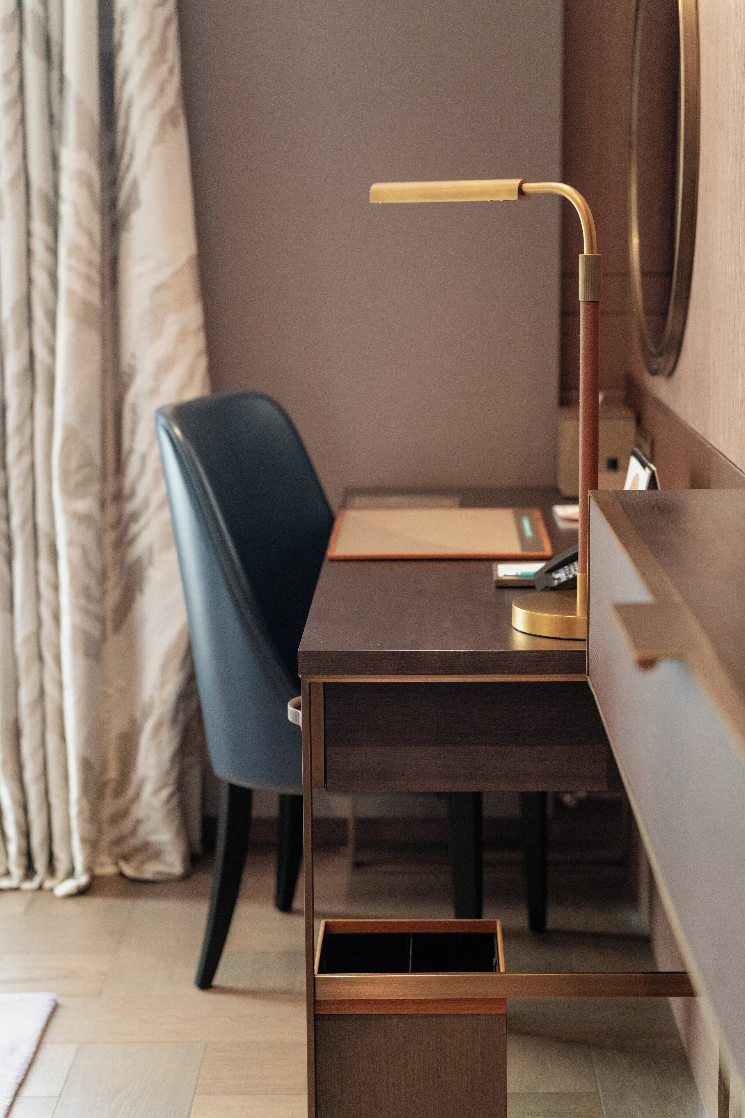 Ein modernes Hotelzimmer mit einem Holztisch, einem blauen Sessel, einer Schreibtischlampe und einem Nachttisch.