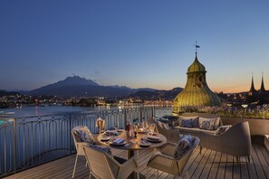 Una terraza con mesa lista para comer, con vista a un cuerpo de agua y un paisaje urbano con montañas al fondo.