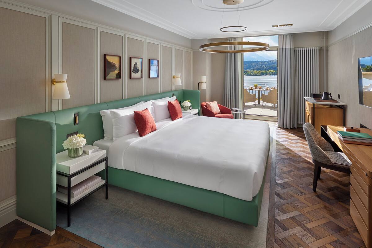 A spacious hotel room with a large bed, a desk with a chair, a view of the outdoors, and a balcony with a railing.