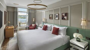 Amplia habitación de hotel con una cama grande, un cabecero tapizado en verde, una mesita de noche con un jarrón de flores, un escritorio con una lámpara y vistas al exterior a través de un ventanal.