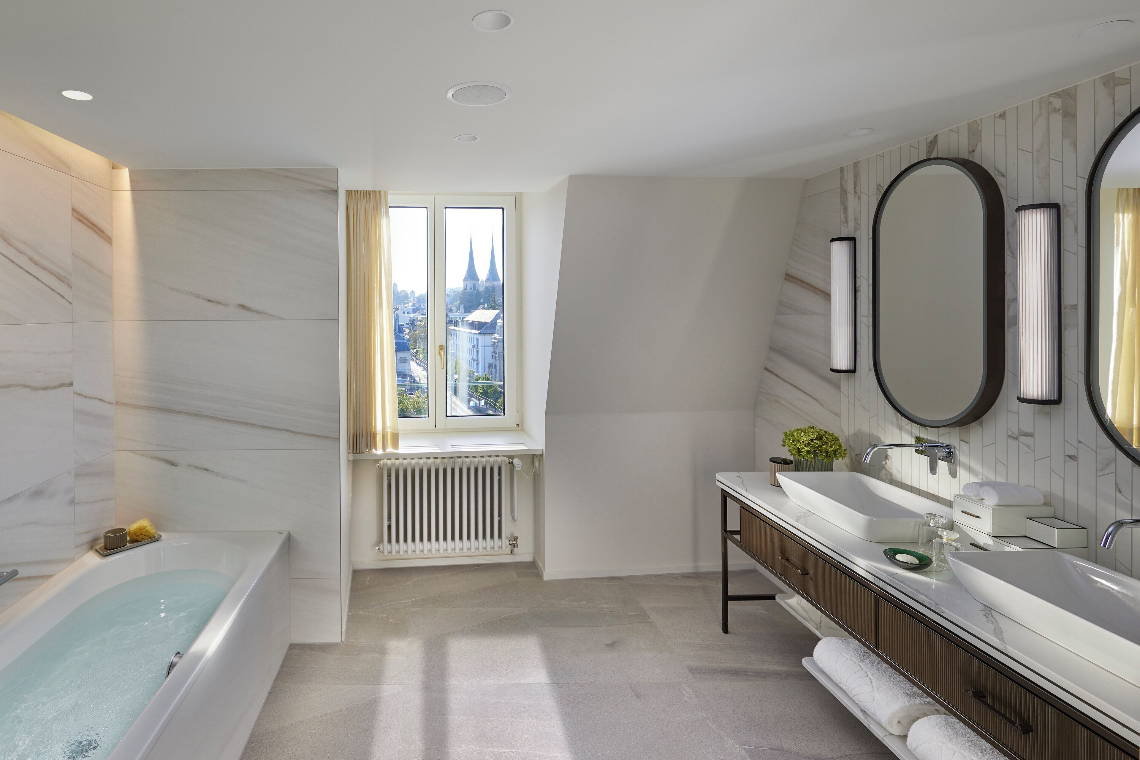 A modern bathroom with a bathtub, a large mirror, and a window offering a city view.