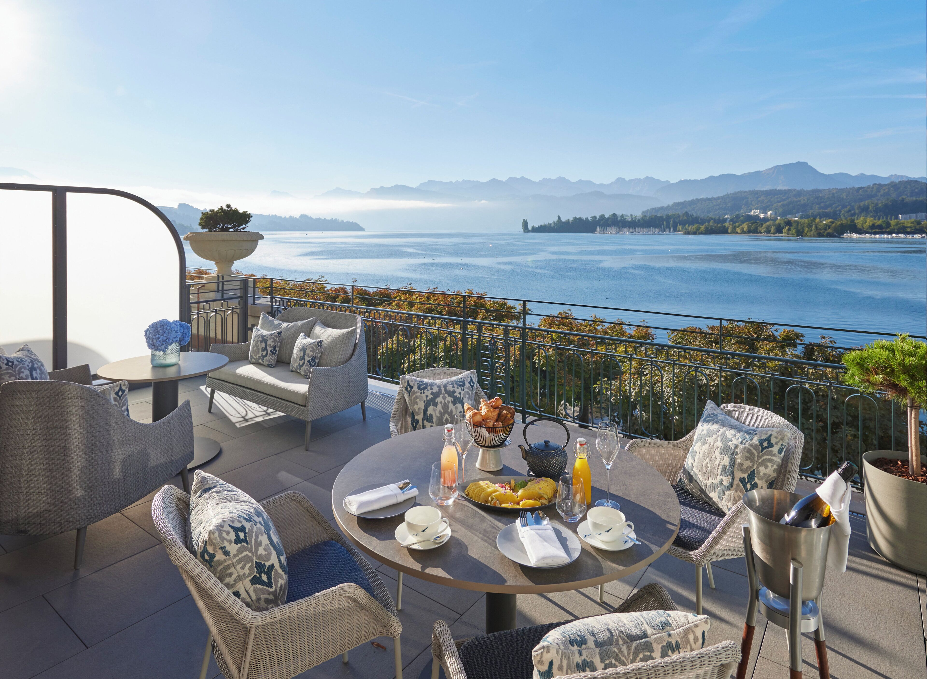 Una terraza con mobiliario exterior, una mesa de comedor preparada para dos personas y vistas a un lago y montañas.