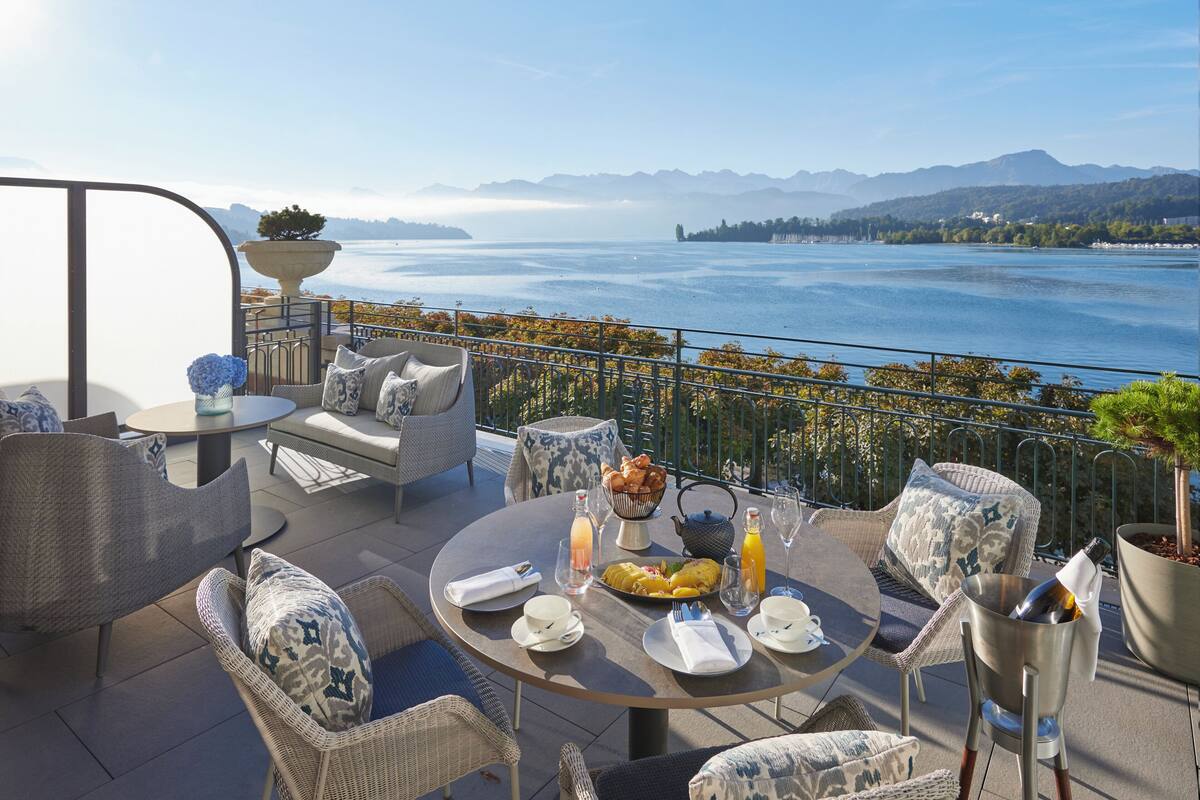 A terrace with wicker furniture, a table set for breakfast, and a view of a lake and mountains.