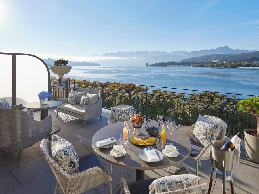 A terrace with outdoor furniture, a dining table set for two, and a view of a lake and mountains.