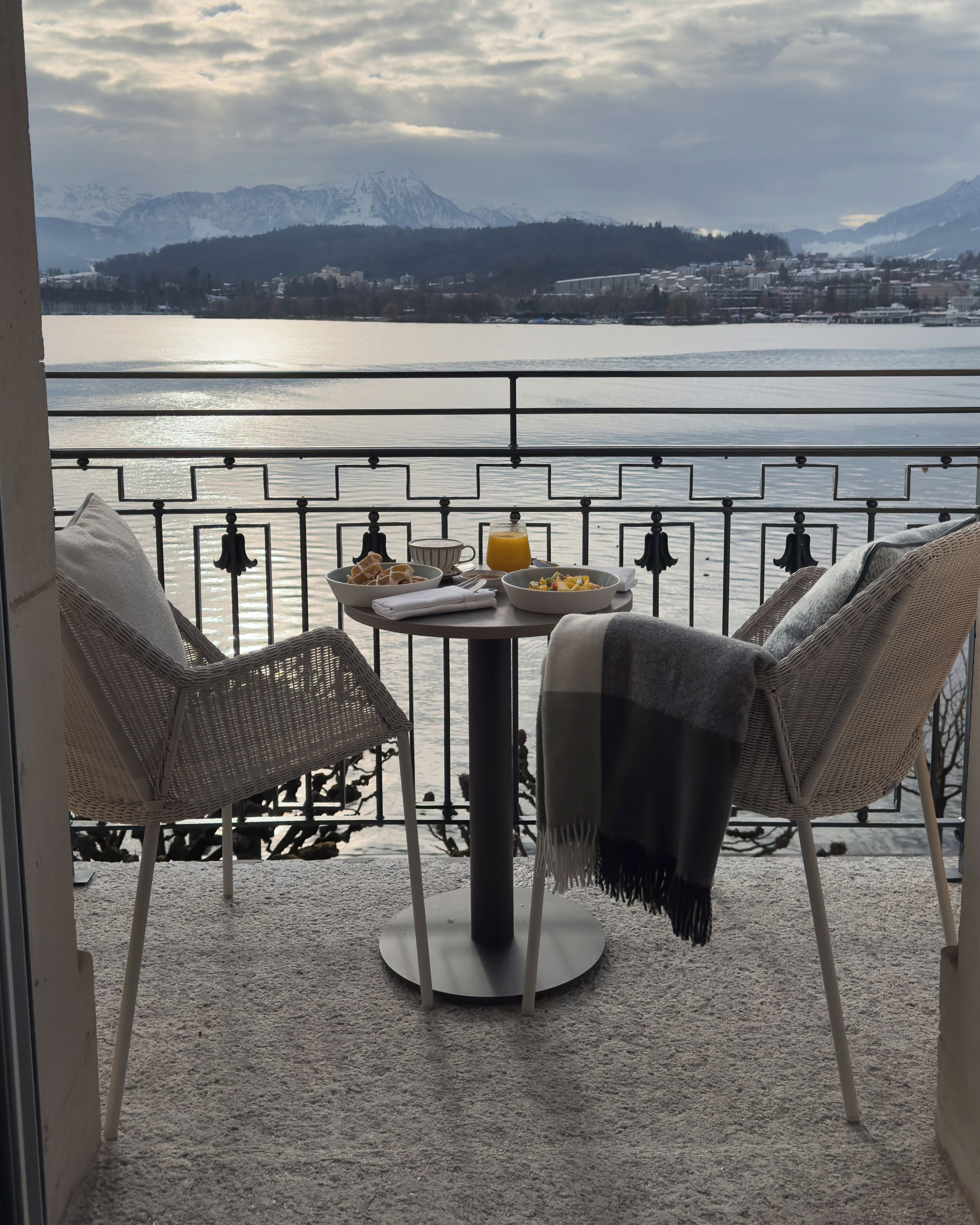 A balcony with a table, two wicker chairs, and a view of a lake and mountains.