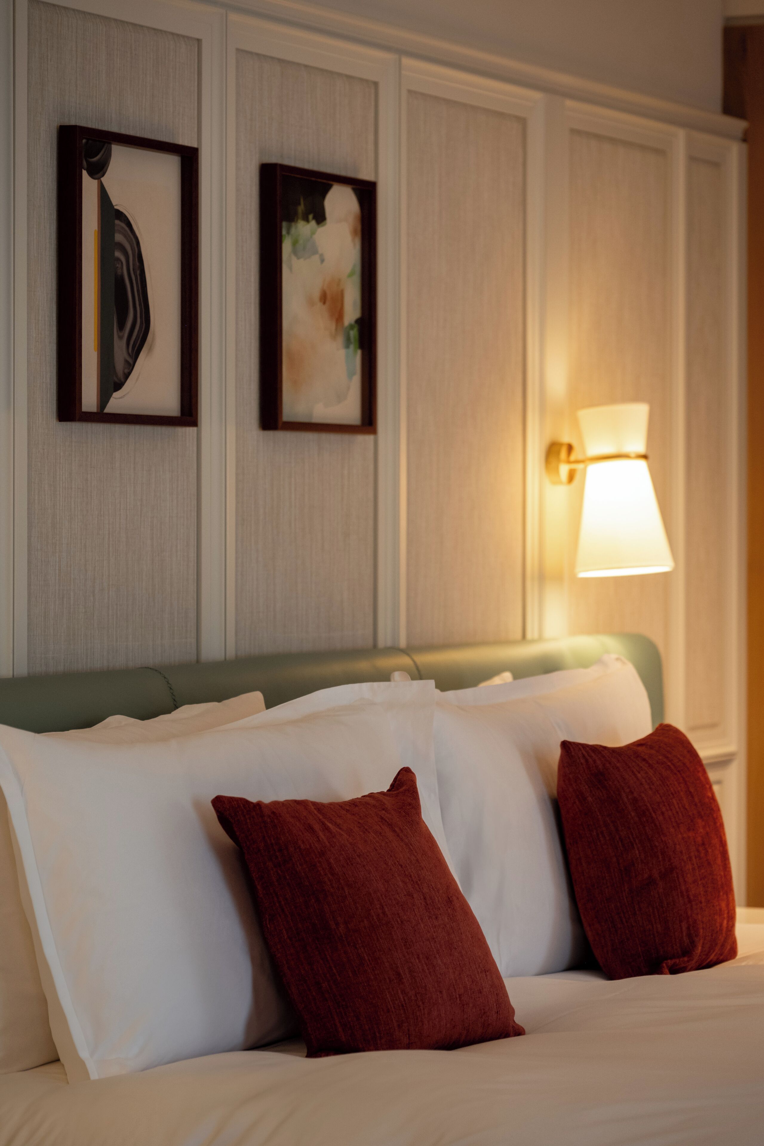 A hotel room with a bed, pillows, a wall-mounted lamp, and two framed artworks.