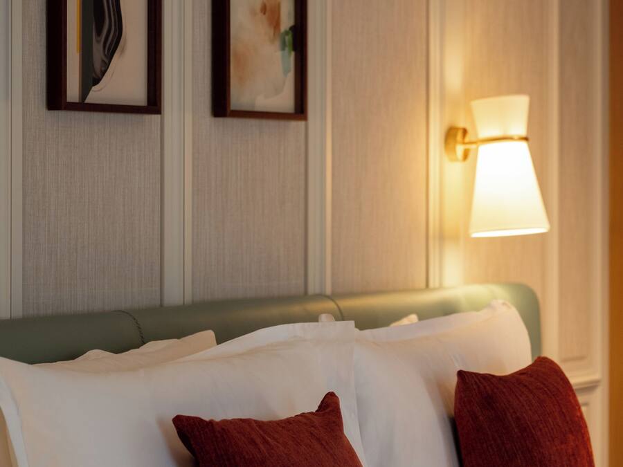 A hotel room with a bed, pillows, a wall-mounted lamp, and two framed artworks.