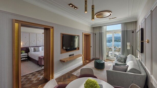 A modern hotel room with a large bed, a TV mounted on the wall, a seating area with a sofa and chairs, and a view of the outdoors.