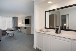 Habitación de hotel con un espejo grande, un lavamanos, una cafetera, un escritorio y un televisor.