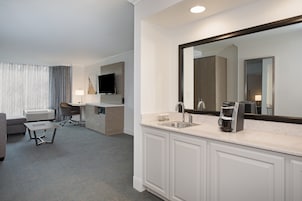 Habitación de hotel con un espejo grande, un lavamanos, una cafetera, un escritorio y un televisor.