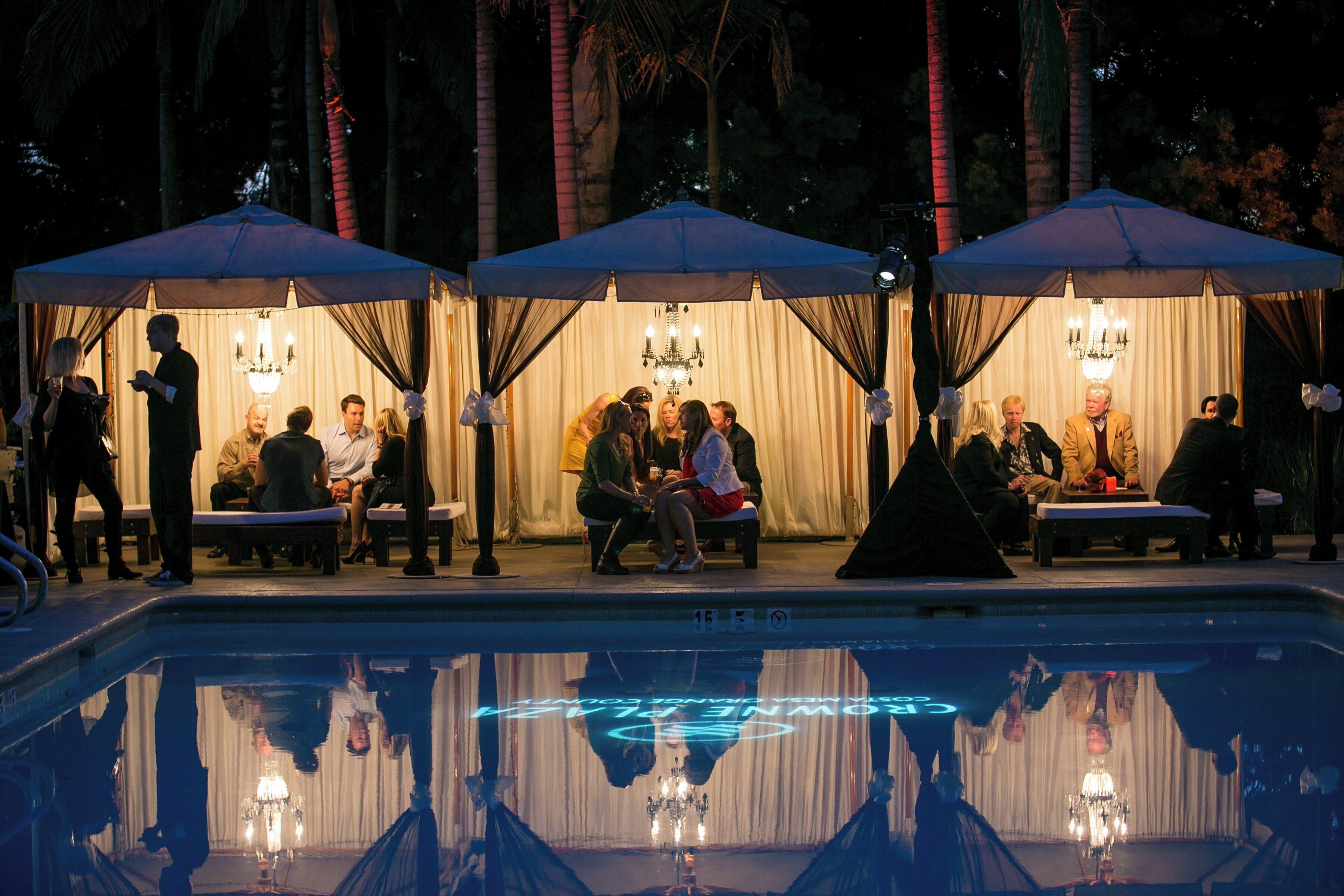 A poolside area with seating under umbrellas, a pool with a reflective surface, and a chandelier.