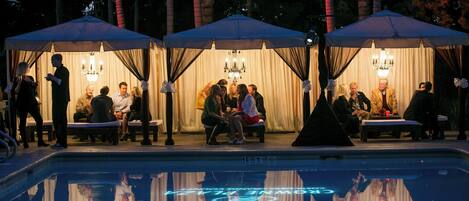 A poolside area with seating under umbrellas, a pool with a reflective surface, and a chandelier.
