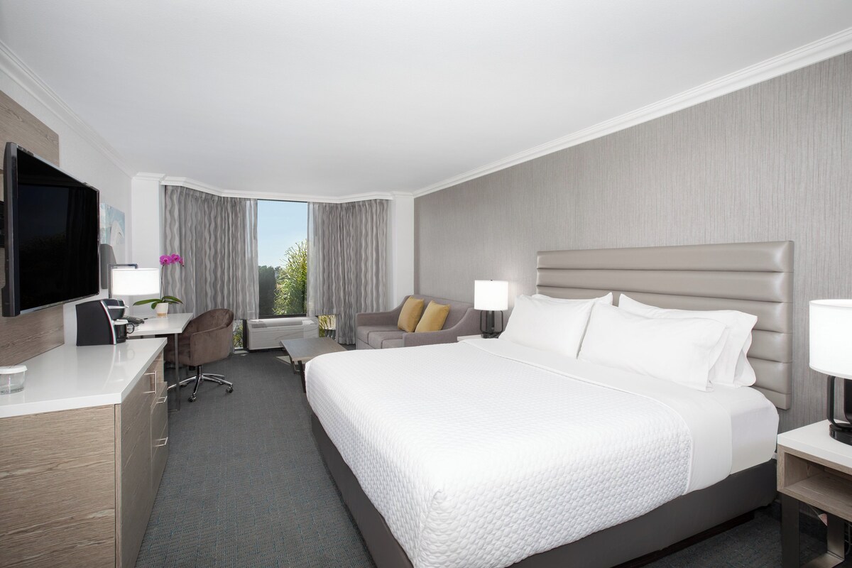 A hotel room with a large bed, a desk with a TV, a chair, and a window with curtains.