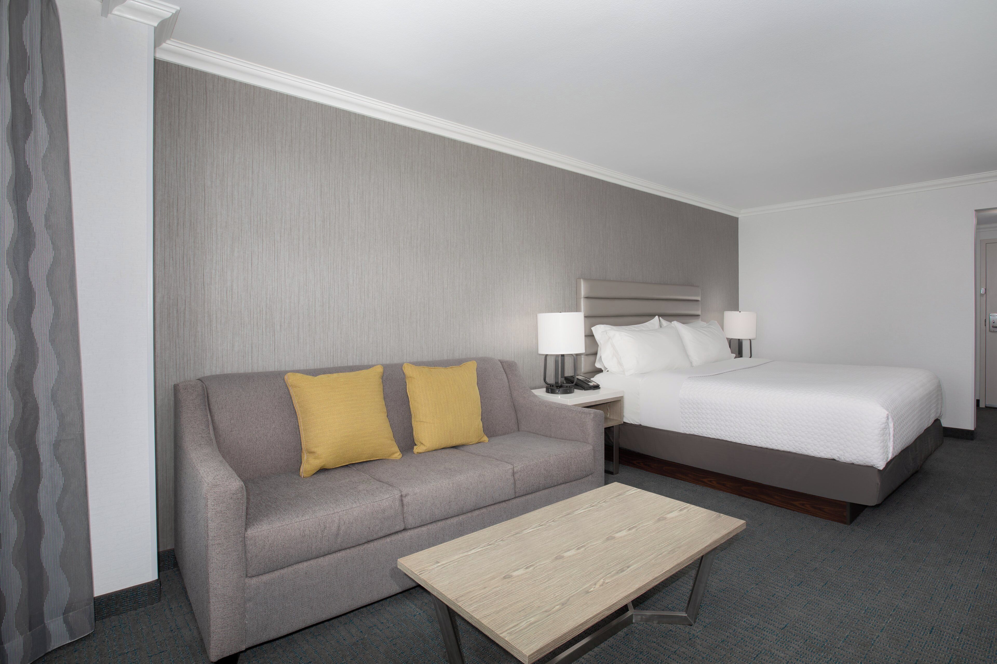 Une chambre d’hôtel avec un canapé gris, un lit, une table de chevet avec des lampes et une petite table en bois.
