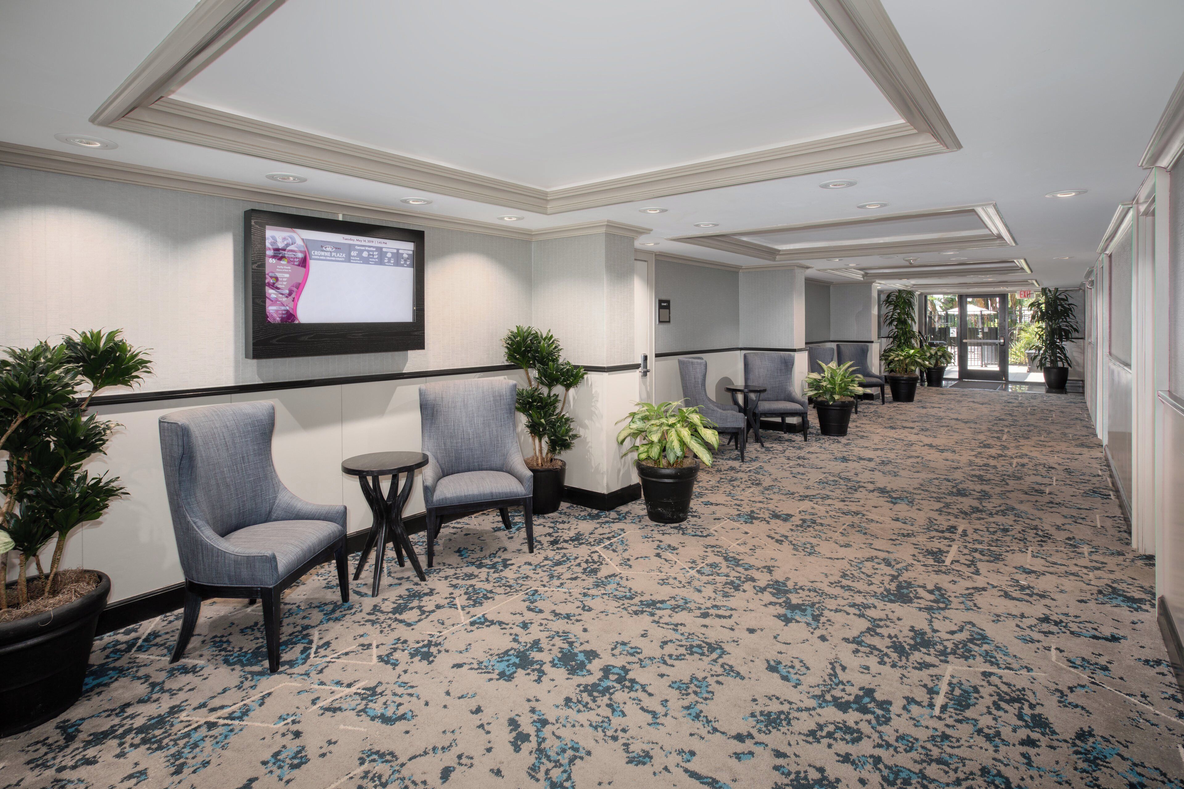 A hotel lobby with a carpeted floor, seating area with chairs, potted plants, and a television screen displaying a weather forecast.