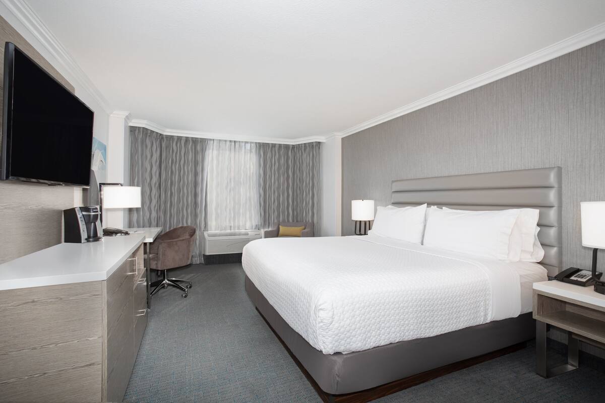A hotel room with a bed, bedside tables, a desk with a chair, a flat-screen TV, and a window with curtains.