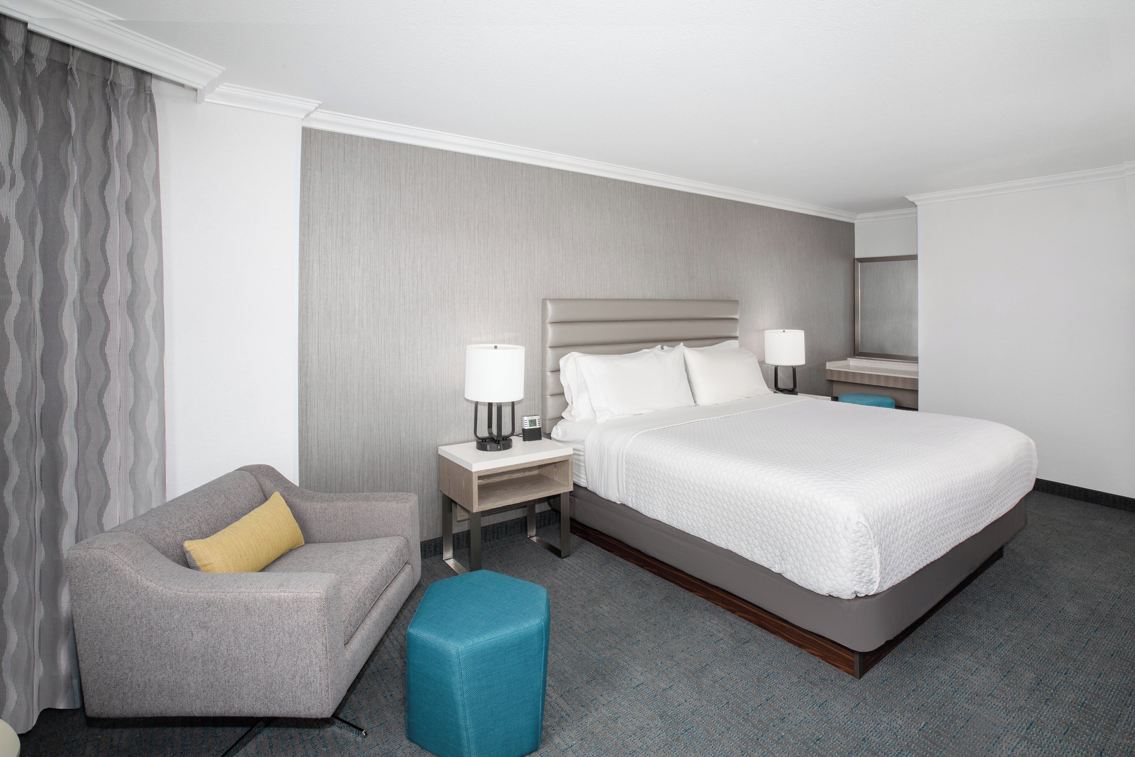 Una habitación de hotel con cama, mesita de noche, lámpara, silla y un pequeño otoman.