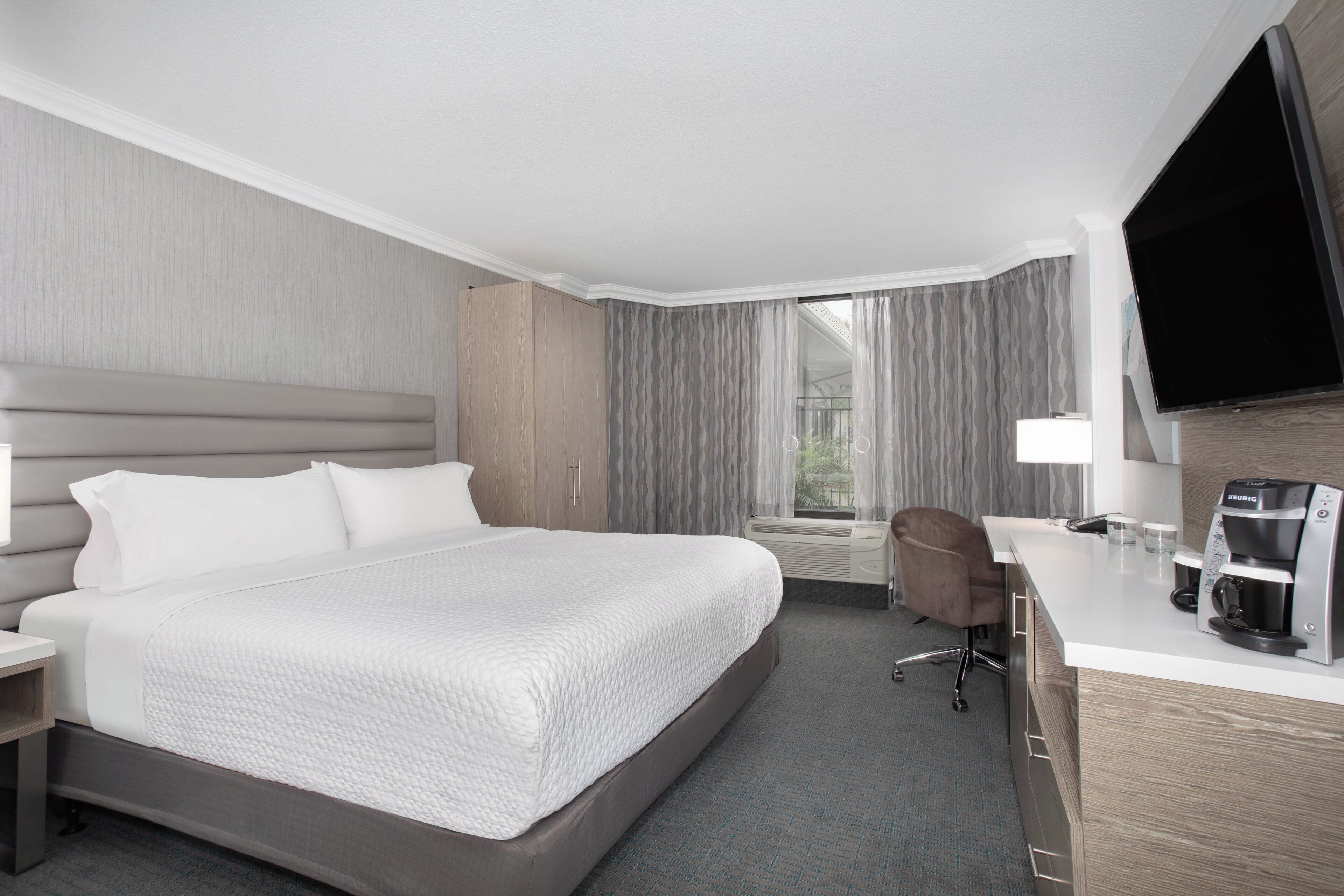 A hotel room with a large bed, a desk with a coffee maker, a TV, and a window with curtains.