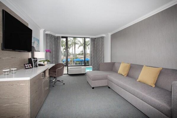 Habitación de hotel con sofá gris, televisor de pantalla plana, escritorio con lámpara y vistas a palmeras a través de la ventana.
