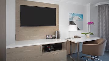 A modern hotel room with a flat-screen TV mounted on a wooden panel, a coffee maker, a microwave, a desk with a chair, and a potted orchid.