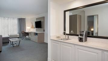 A hotel room with a large mirror, a desk, a chair, a sofa, a coffee maker, and a television.