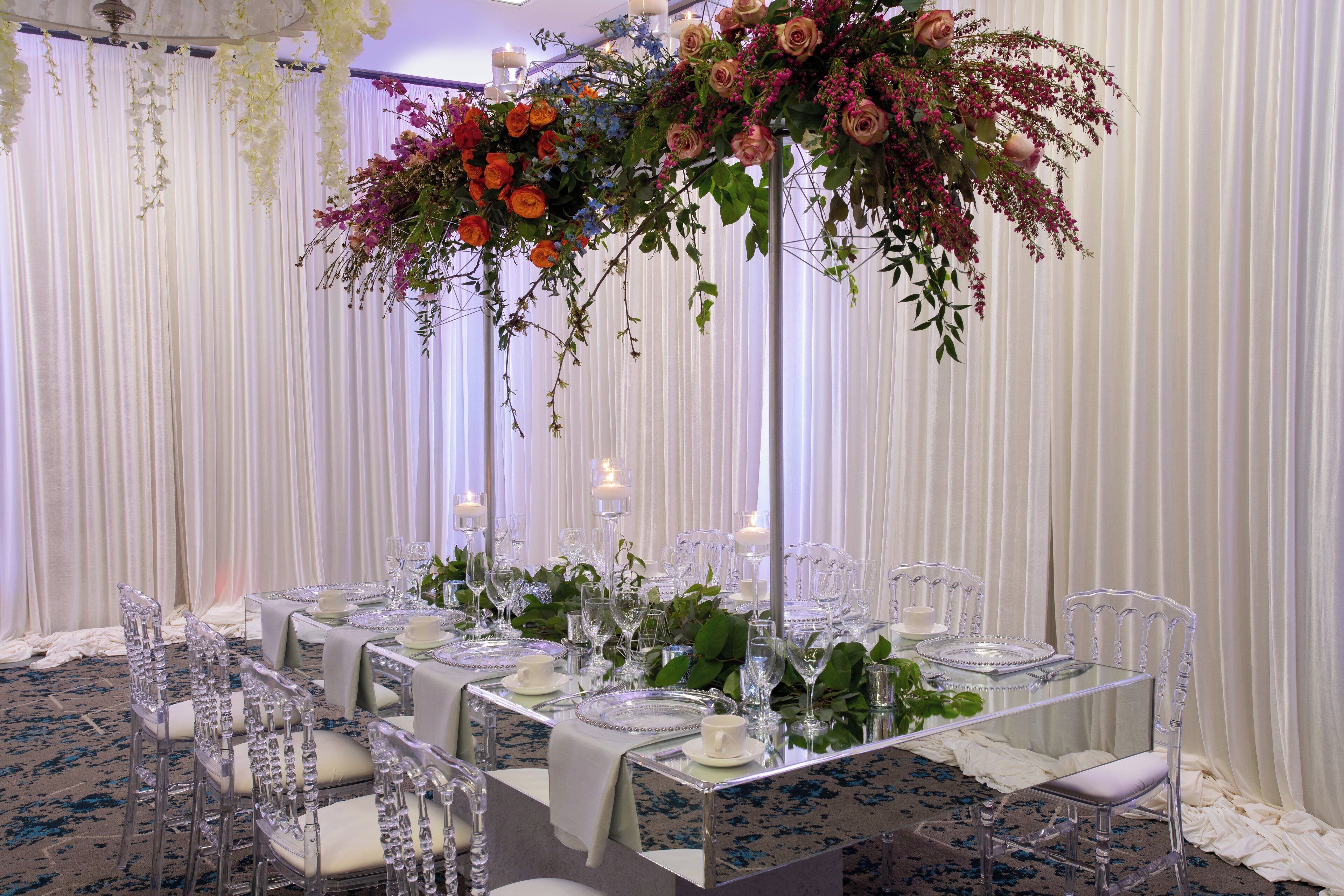 Ein festlich gedeckter Tisch mit klarem Glas und weißen Tischdecken, umgeben von klaren Stühlen, in einem Raum mit weißen Vorhängen und floralen Dekorationen.