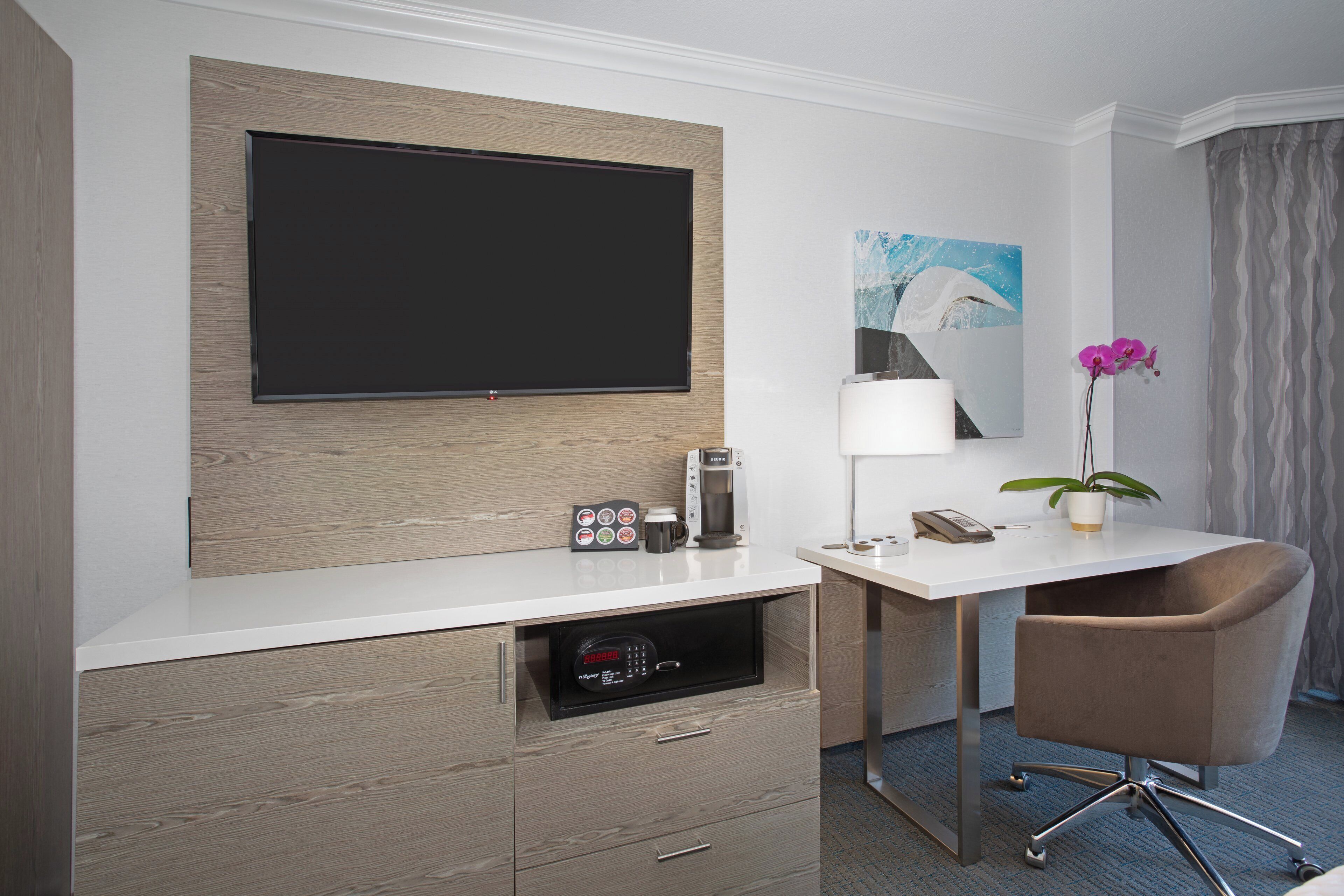 A hotel room with a flat-screen TV, a coffee machine, a desk with a chair, and a potted orchid.