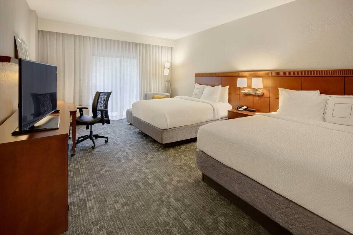 Une chambre d’hôtel avec un grand lit, un plus petit lit, un bureau avec une télévision, une chaise et une fenêtre avec des rideaux.