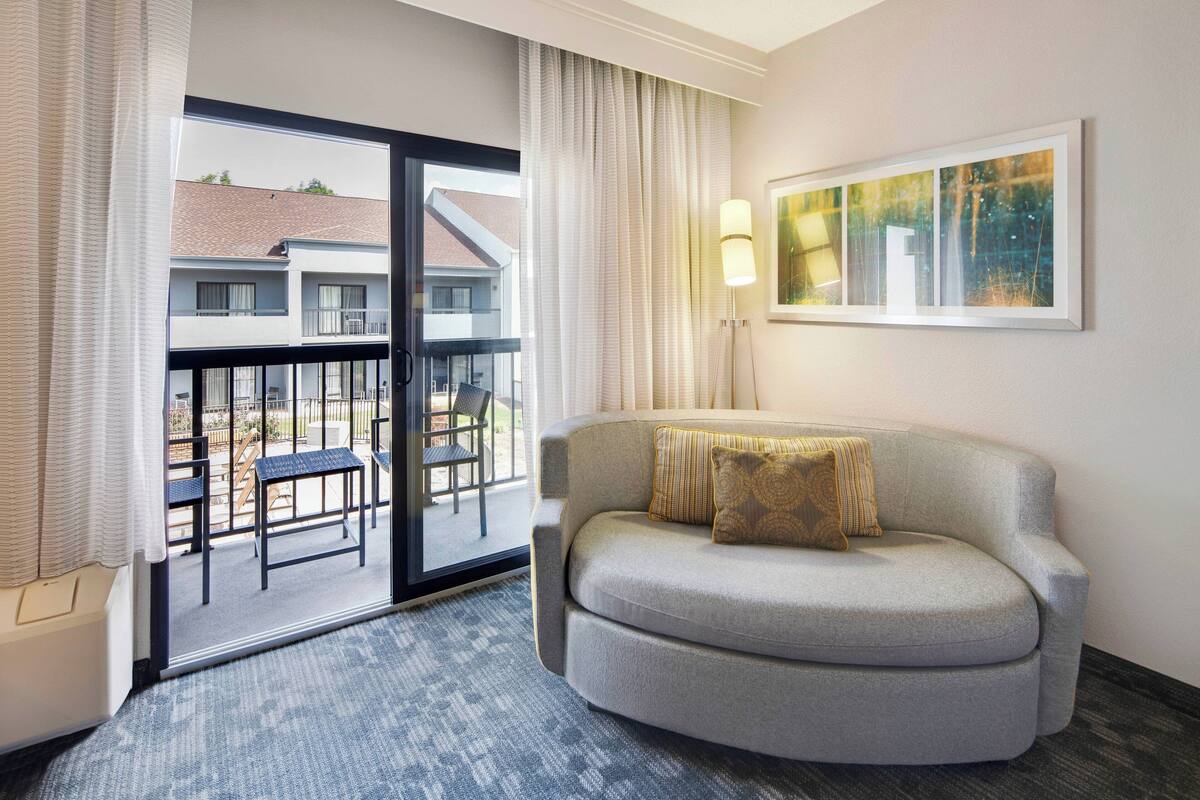 Une chambre d’hôtel avec un canapé gris, une grande fenêtre avec des rideaux, un lampadaire et un tableau au mur.
