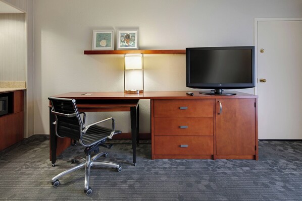 A hotel room with a wooden desk, a black office chair, a flat-screen TV, a lamp, and a door.
