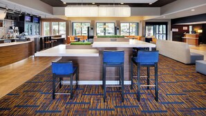 A modern hotel lobby with a central bar area, seating, and a reception desk.