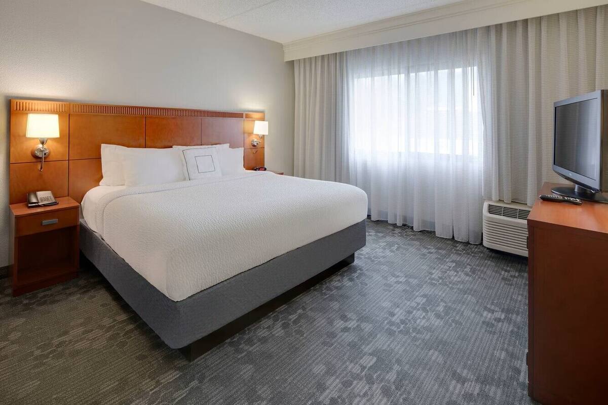 Une chambre d’hôtel avec un grand lit, deux tables de chevet avec des lampes, une télévision et une fenêtre avec des rideaux.
