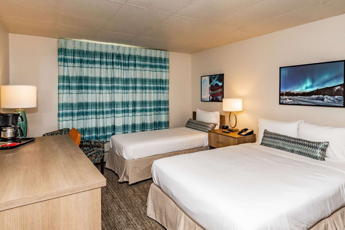 A hotel room with two beds, a desk, a chair, a coffee maker, and a nightstand with a lamp.