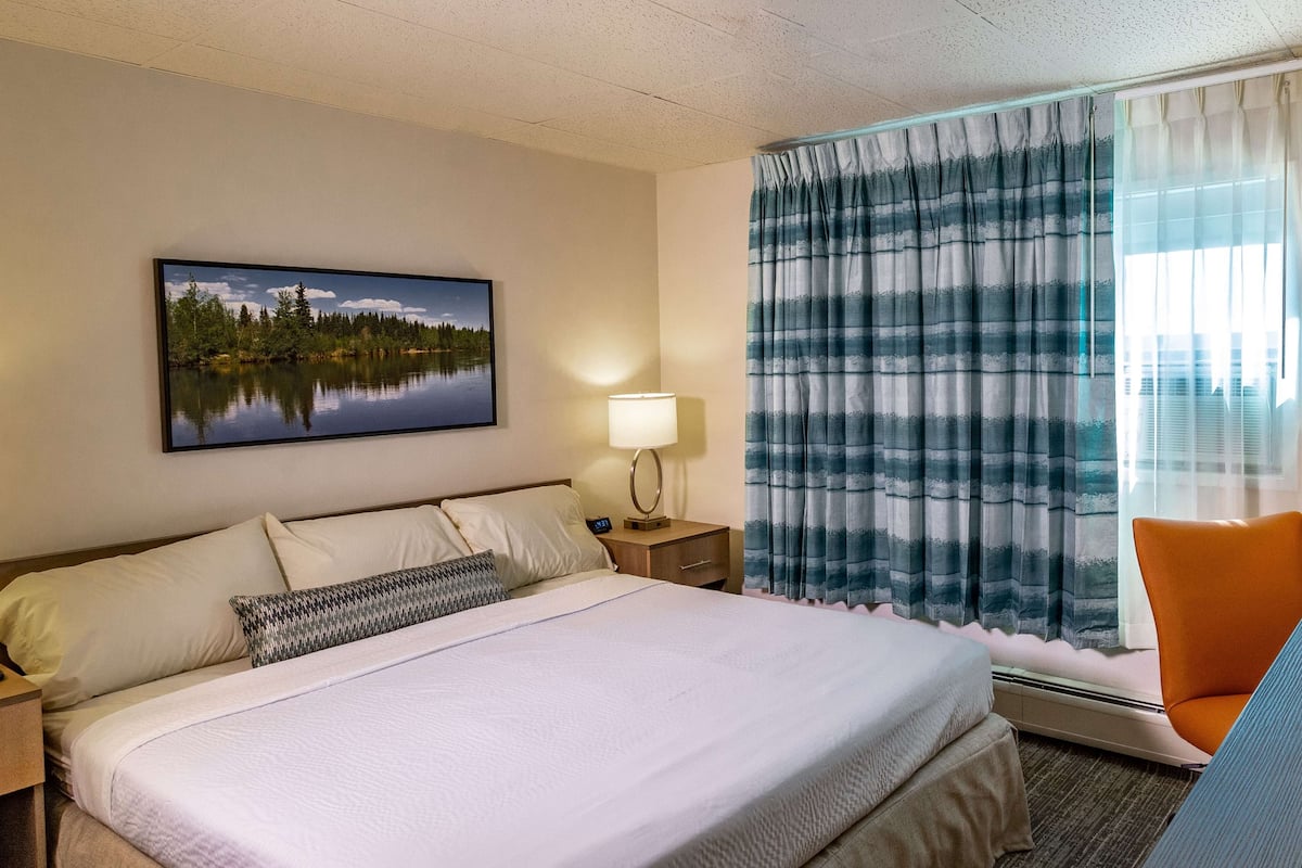 A hotel room with a bed, a nightstand with a lamp, a window with curtains, and a chair.