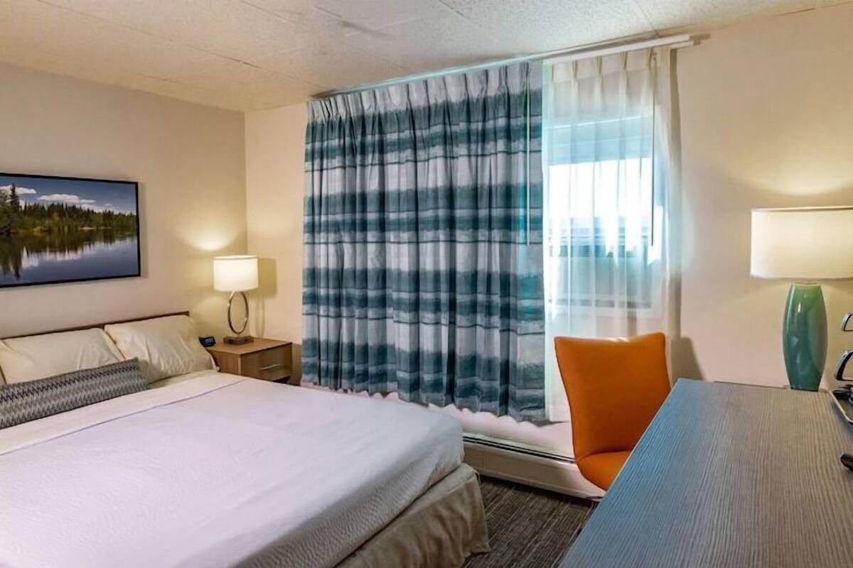 A hotel room with a bed, bedside lamps, a desk with a chair, a window with curtains, and a painting on the wall.