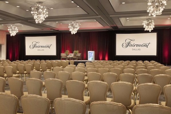 A conference room with rows of beige chairs, a stage with a screen displaying "Fairmont Dallas," and chandeliers.