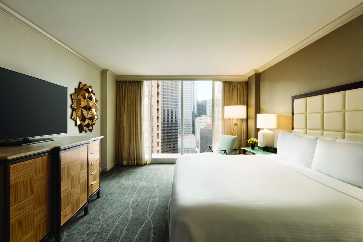 A hotel room with a large bed, a TV on a wooden cabinet, a city view through the window, and a small table with a lamp.