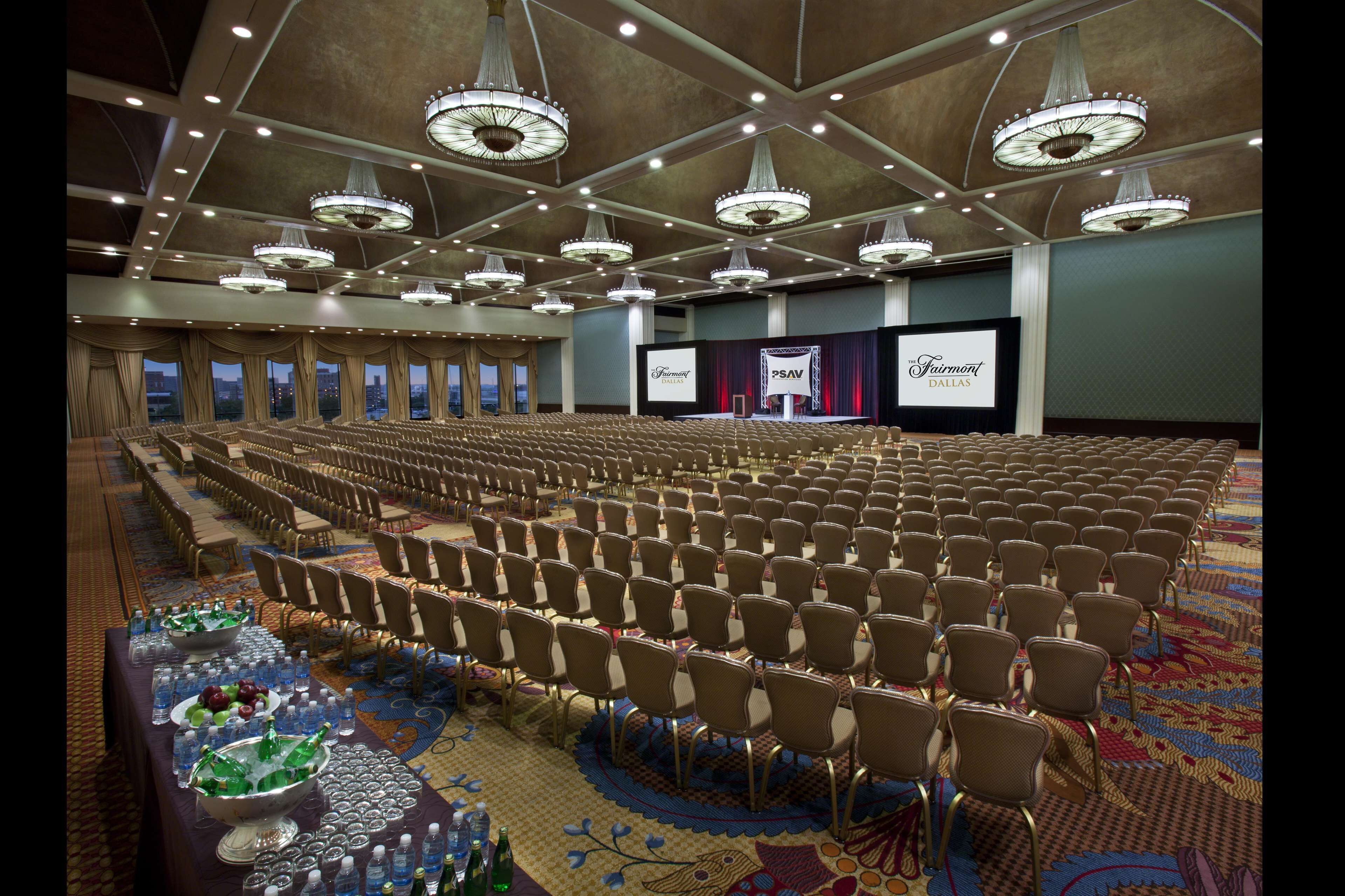A large conference room with rows of chairs, a stage with a screen, and a buffet table with drinks and snacks.