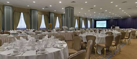 Un salón de banquetes con mesas redondas dispuestas para cenar, un escenario con una pantalla y suelo alfombrado.