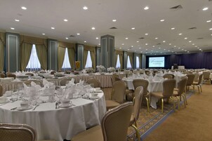Un salón de banquetes con mesas redondas dispuestas para cenar, un escenario con una pantalla y suelo alfombrado.