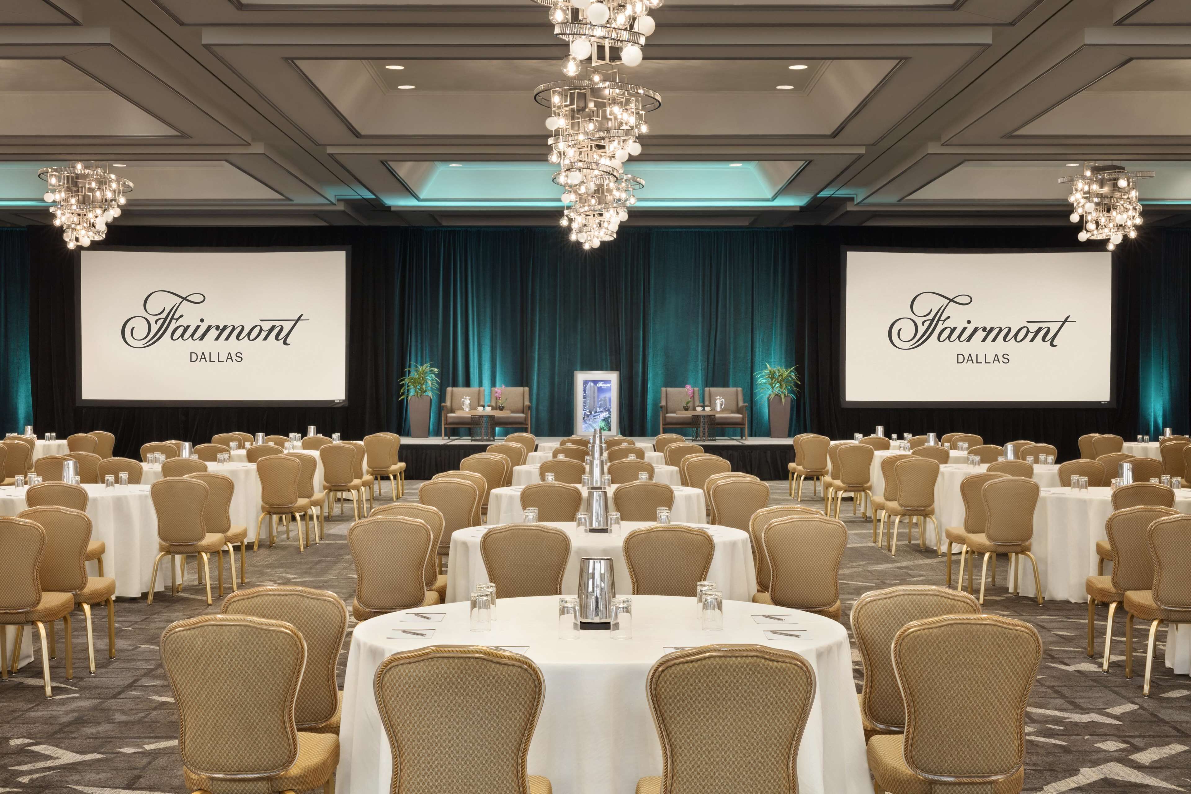 A conference room with round tables and chairs, a large screen displaying 'Fairmont Dallas', and a chandelier.