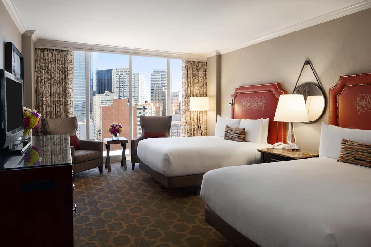 A hotel room with two beds, a desk with a TV, a chair, a small table with flowers, and a view of the city skyline.