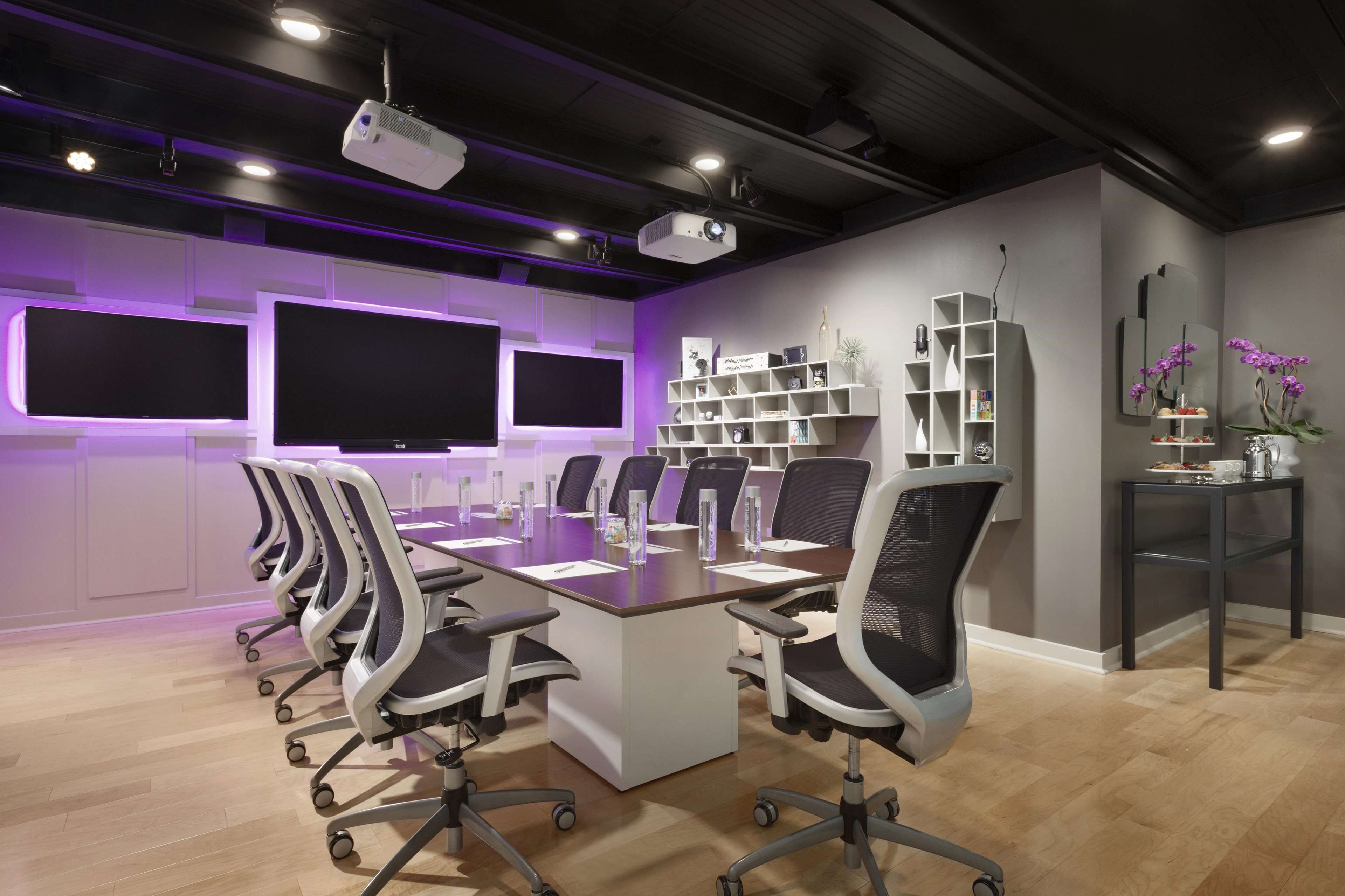 Un salón de conferencias con una larga mesa, varias sillas, un proyector y estanterías con libros y objetos decorativos.